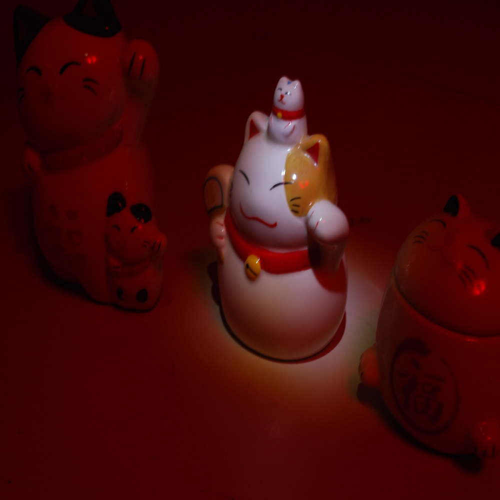 image of 3 porcelain lucky cats in red light, with one cat being illuminated in the center by white light